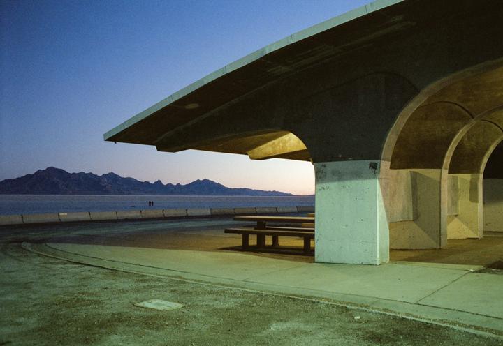 A concrete structure with picnic tables sits beside a body of water with mountains in the distance under a twilight sky.