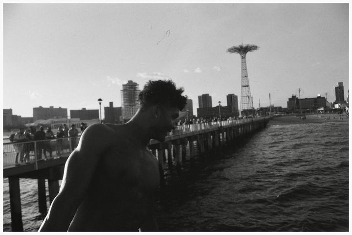 A shirtless man stands on a pier with a city skyline and a parachute jump tower in the background.