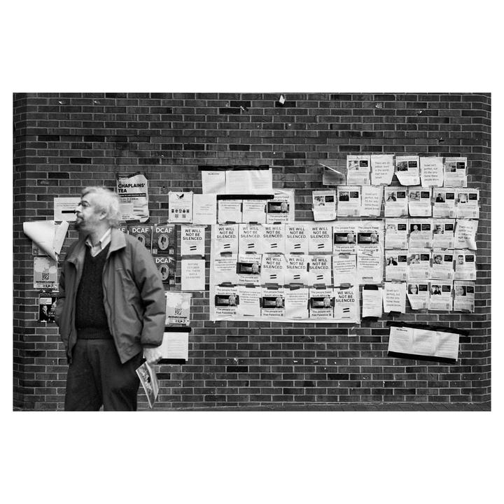 A man stands next to a brick wall covered in flyers and posters.