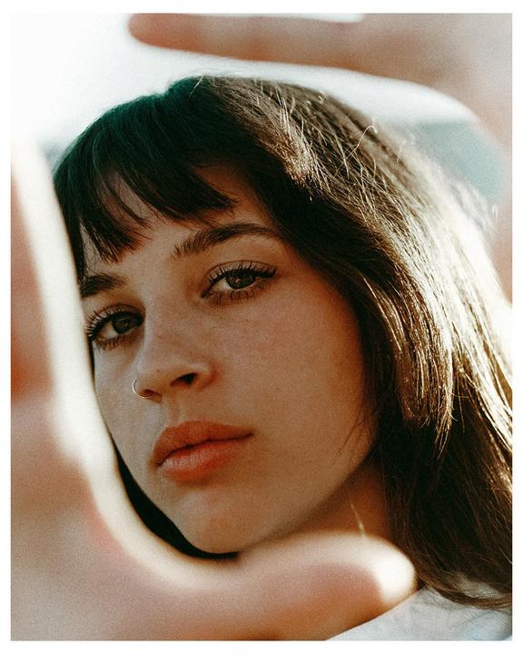 A woman with bangs and a nose ring looks directly at the viewer, framed by hands.