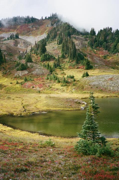 A small lake sits at the base of a mountain covered in trees and colorful autumn foliage under a hazy sky.