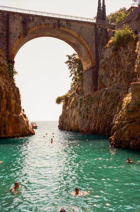 A stone bridge arches over a narrow inlet where people are swimming in turquoise water.