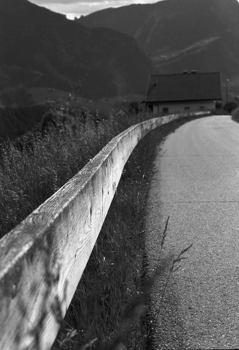 A wooden fence runs alongside a paved road leading towards a house and mountains in the distance.