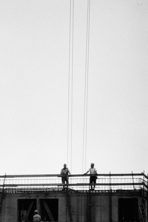 Two men stand on a building under construction, with a third man visible below them.