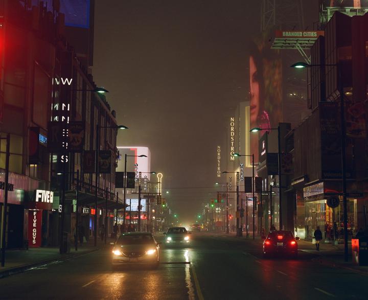 A foggy street at night is illuminated by neon signs and car headlights.