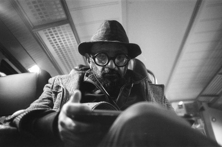 A man wearing a hat and glasses looks directly at the viewer while holding a phone.