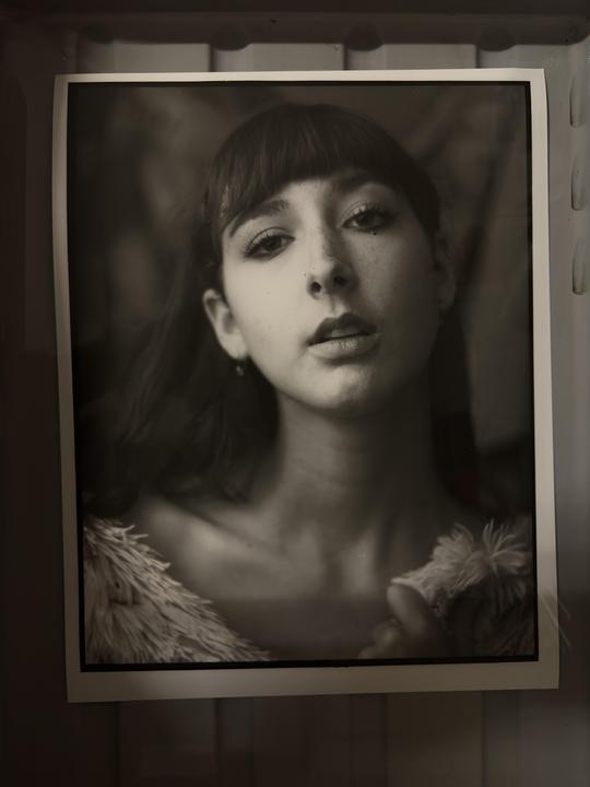 A woman with bangs and a fluffy garment looks directly at the viewer in a black and white portrait.