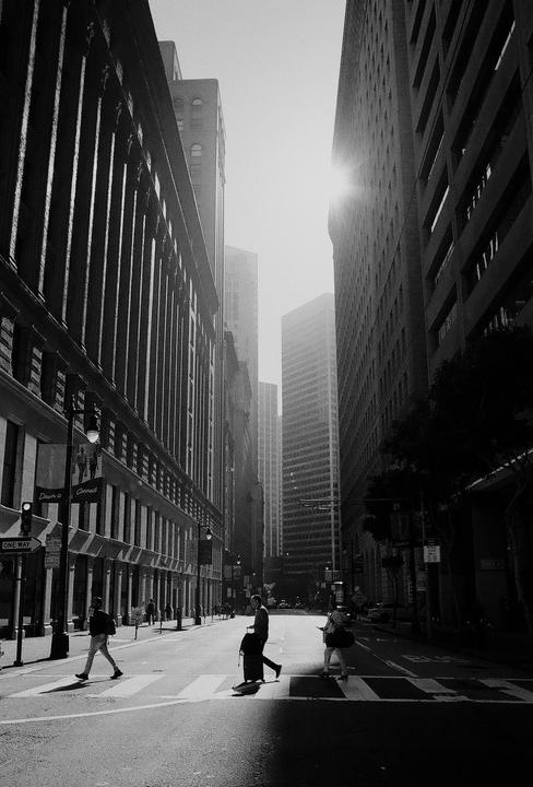Three people cross a street in a city with tall buildings and bright sunlight.