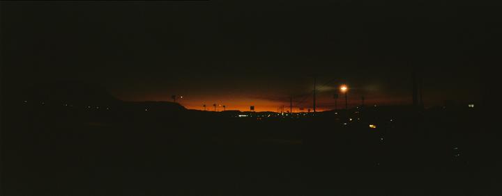 A panoramic view of a city at dusk shows a bright orange sky above a dark landscape with scattered lights.