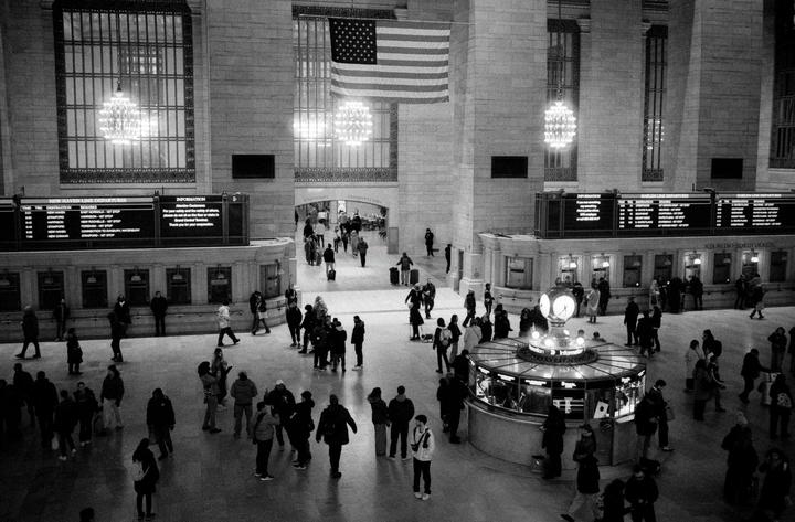 A large crowd of people are gathered in a grand hall with an American flag hanging from the ceiling.