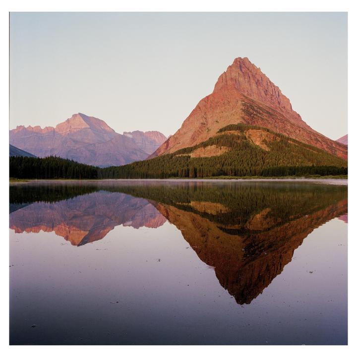 A large mountain with trees on its lower slopes is reflected in a still lake under a clear sky.