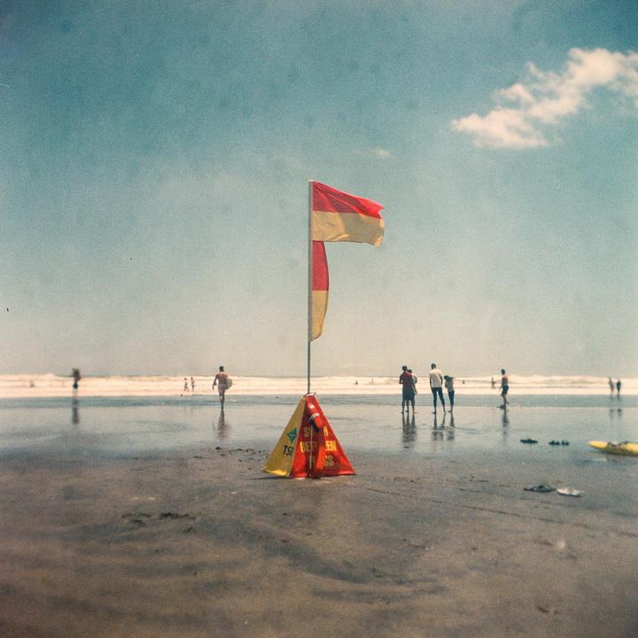 A red and yellow flag stands on a wet beach with people and waves in the background.