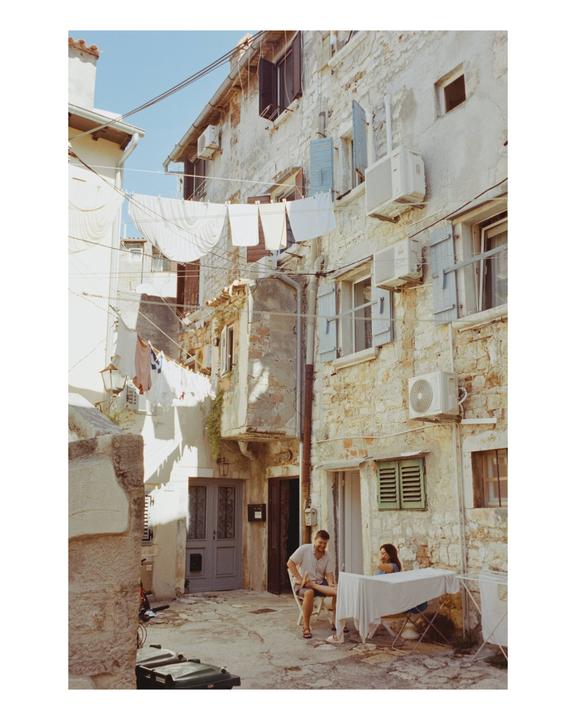 A man and a woman sit at a table in a narrow alleyway with laundry hanging overhead.