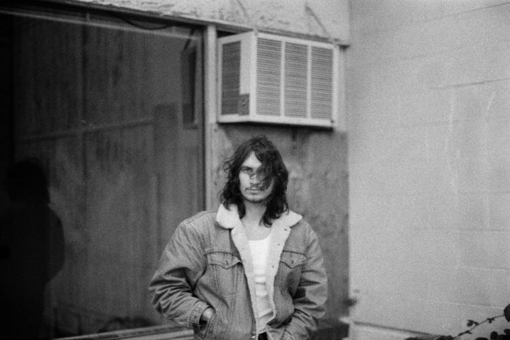 A man with long hair and a denim jacket stands in front of a building with an air conditioner.