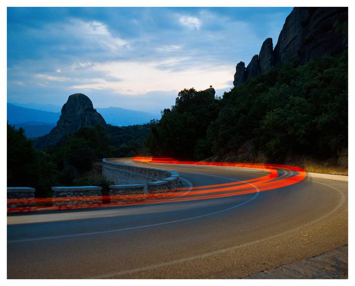 A winding road with red light trails curves through a mountainous landscape at dusk.