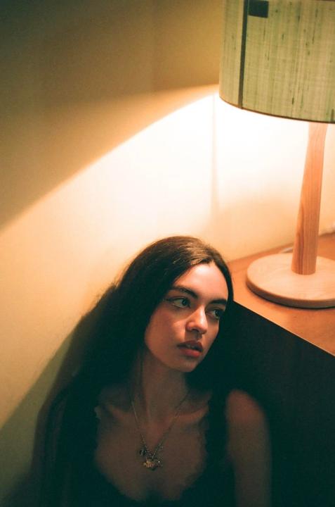 A woman with long dark hair sits on the floor next to a lamp, looking away from the camera.