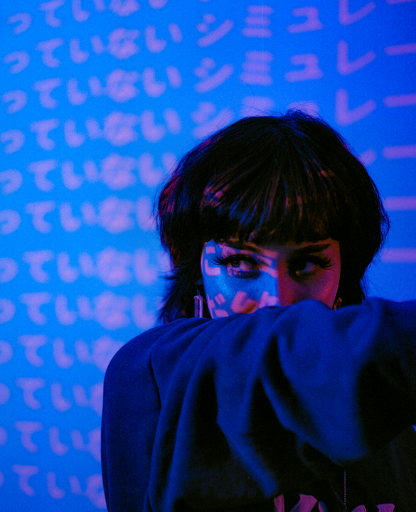 A woman with dark hair looks to the side, with Japanese characters projected onto her face and the wall behind her.