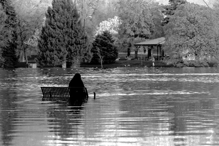 A person sits on a partially submerged park bench in a flooded park with trees and a gazebo in the background.