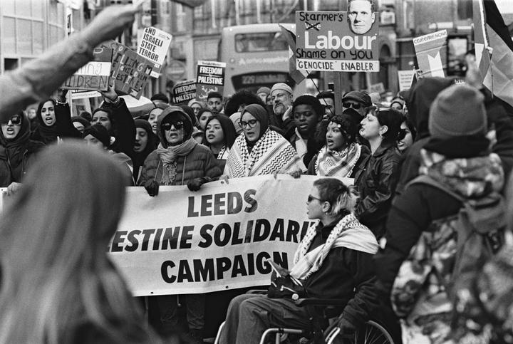 A crowd of people protest in a street, holding signs and a banner.