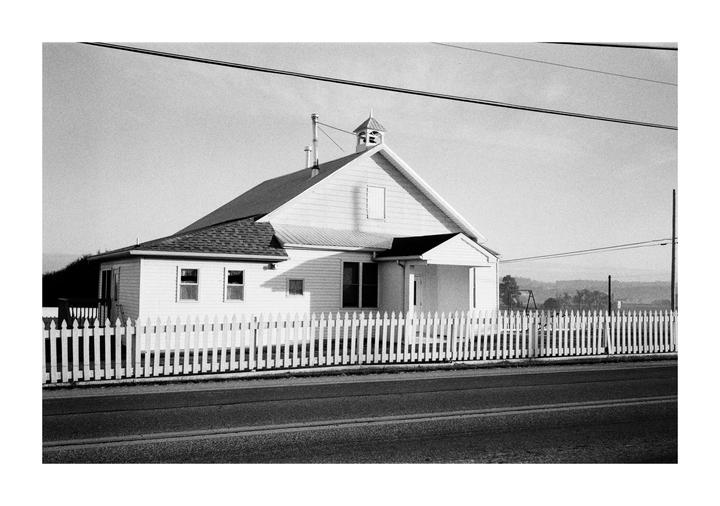 A white house with a steeple and a picket fence stands beside a road under a clear sky.