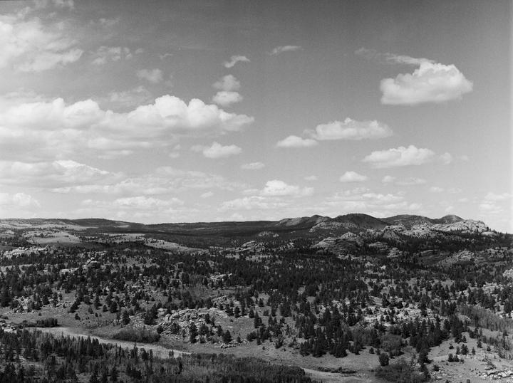 A vast landscape of rolling hills and mountains covered in trees and rocks stretches under a sky with scattered clouds.