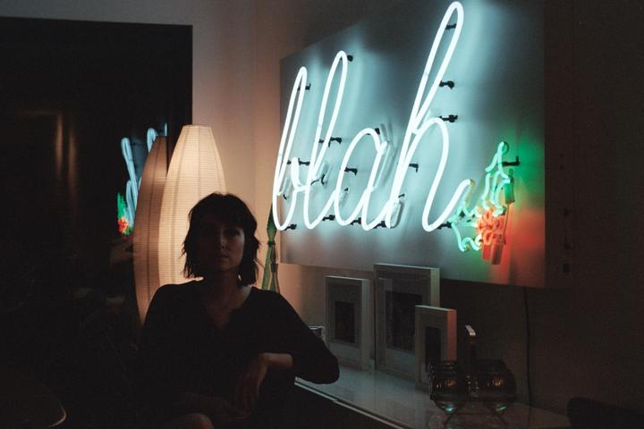 A woman sits in a dimly lit room with a neon sign that reads "blah" on the wall.