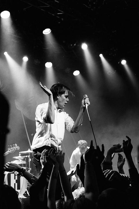 A man in a white shirt sings into a microphone on stage, with many hands raised in the foreground and spotlights shining down.