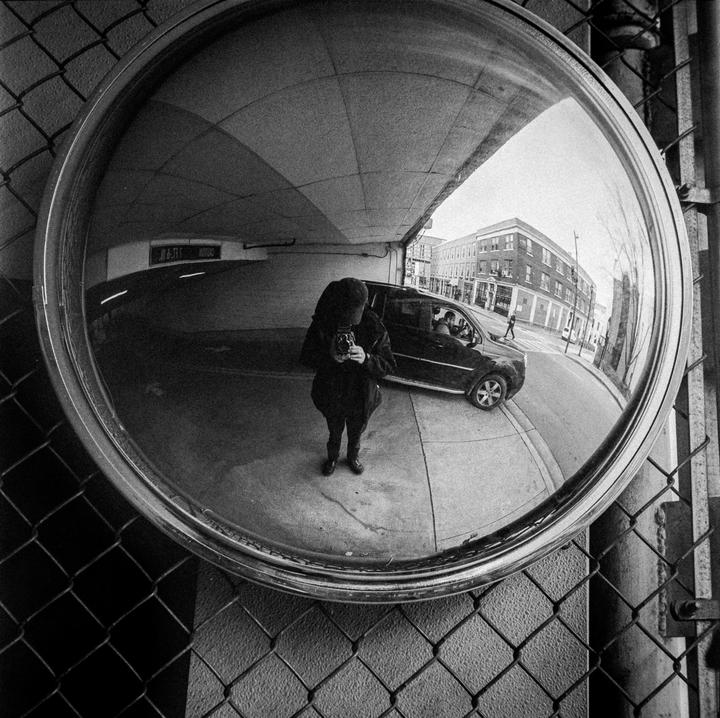 A person holding a camera is reflected in a convex mirror, with a street and building visible in the background.