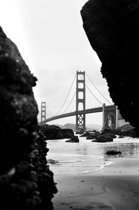 The Golden Gate Bridge is visible in the distance through a gap between two large rocks on a beach at low tide.