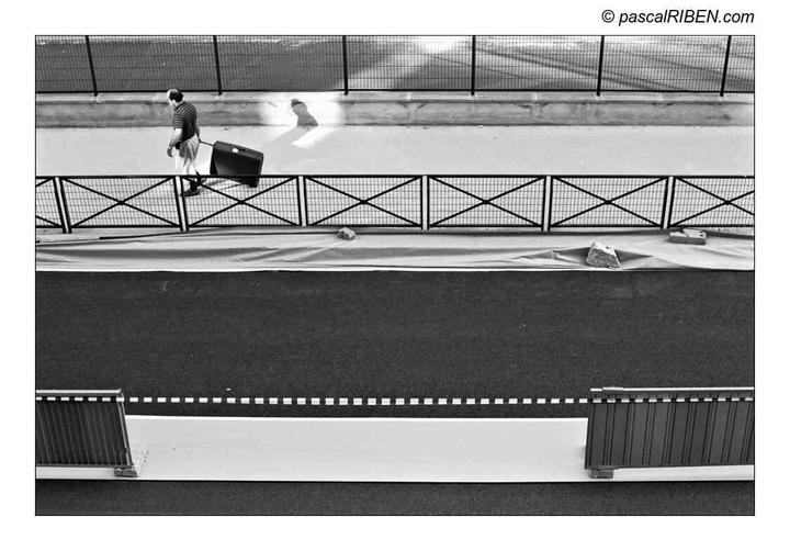 A man pulls a suitcase along a street next to a fence.