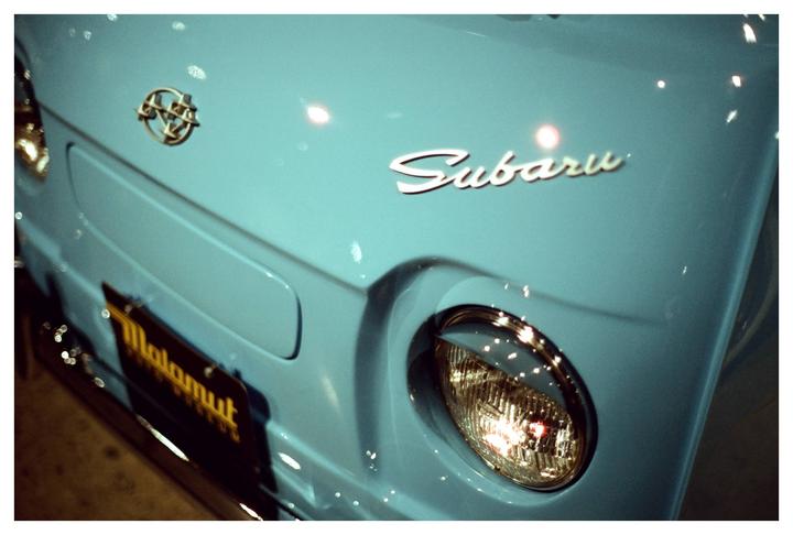 The front of a light blue Subaru car is visible, showing its emblem, name, and a headlight.