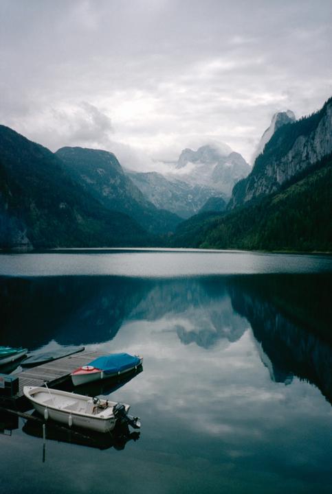 A calm lake reflects mountains and an overcast sky, with several boats docked at a wooden pier in the foreground.