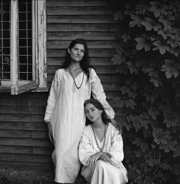 A woman stands next to a girl who is kneeling, both wearing long dresses and necklaces, in front of a wooden building with a window and vines.