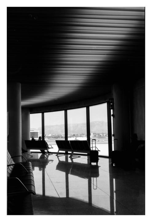 Two people sit on benches in an airport terminal with large windows overlooking a city and mountains.