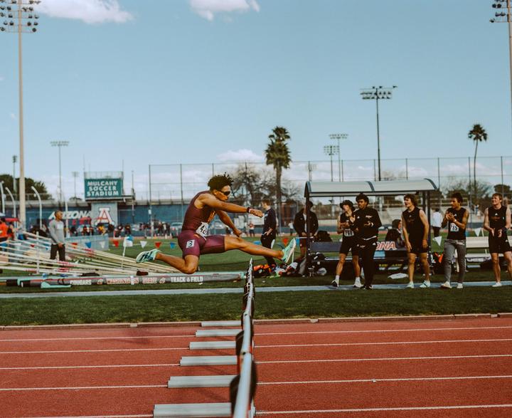 A man in athletic wear jumps over a hurdle on a track and field course.