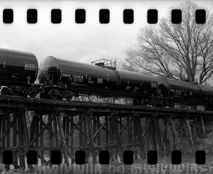 A black and white photograph shows a train with several tanker cars crossing a wooden trestle bridge over water, with a bare tree in the background.