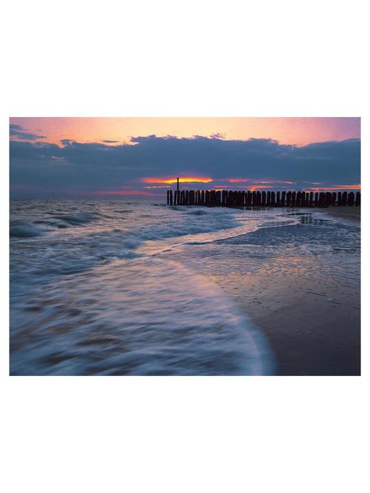 Waves crash on a beach next to a wooden pier at sunset.