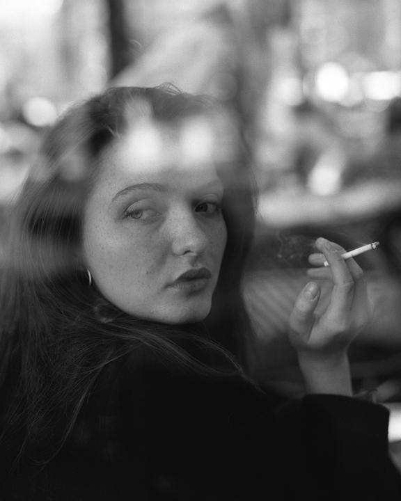 A woman with long hair and freckles looks over her shoulder while holding a lit cigarette, with reflections visible on a window in the foreground.