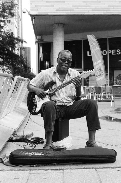 A man wearing sunglasses and a plaid shirt sits on a speaker and plays an electric guitar on a city street.