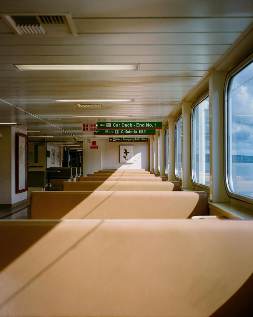 A long row of empty seats on a ferry boat is illuminated by strong diagonal sunlight from the windows.
