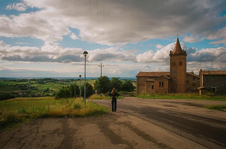 A woman stands on a road looking out at a landscape with a church and rolling hills under a cloudy sky.