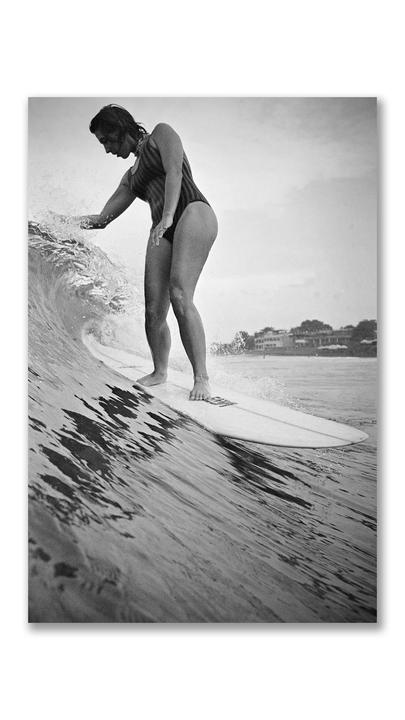 A woman in a striped swimsuit surfs a wave, with buildings visible on the shore in the background.