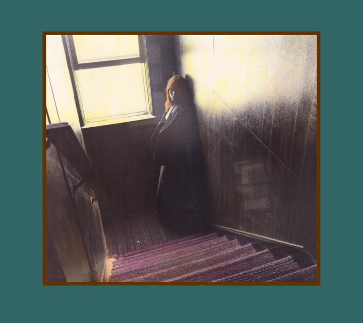 A woman stands in a dark stairwell next to a window, with light illuminating the wall behind her.