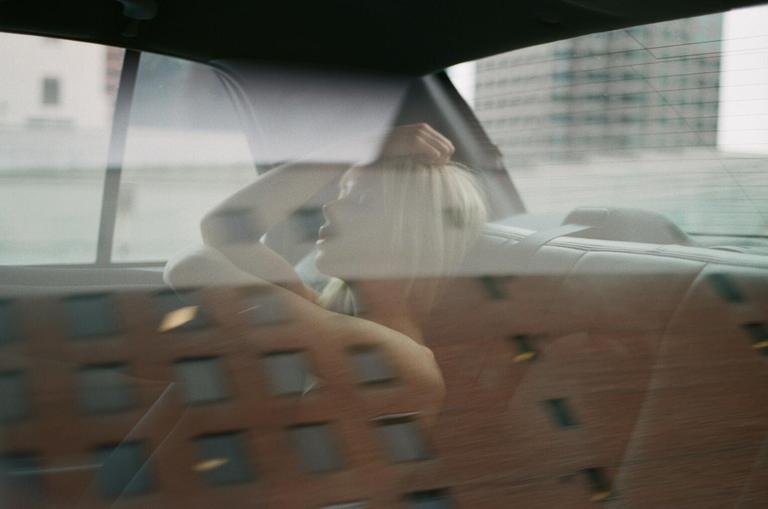 A woman with blonde hair is visible through a car window, with reflections of buildings superimposed on the image.
