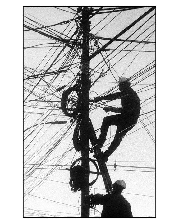 Two men are working on a utility pole with many wires against a bright sky.