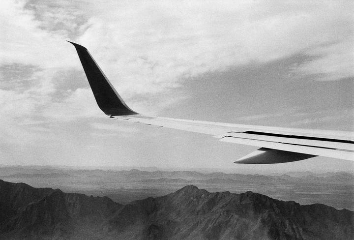 An airplane wing is visible above a mountain range under a cloudy sky.