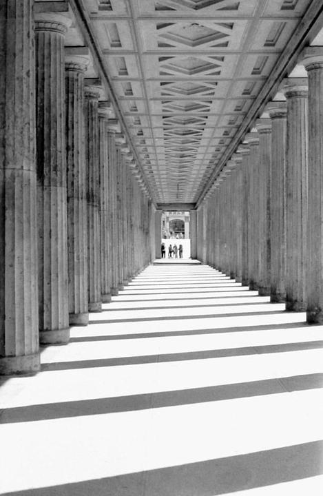 A long corridor with columns and a patterned ceiling leads to a group of people in the distance, with strong shadows cast across the floor.