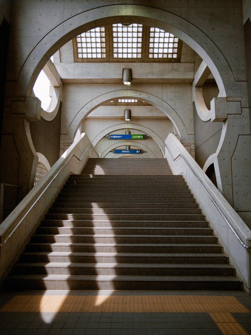 A long concrete staircase with arched ceilings and windows leads upwards, with shadows cast across the steps.