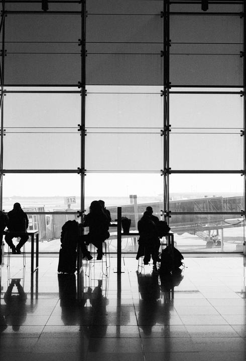Silhouetted people sit at tables in front of a large window overlooking an airport tarmac.