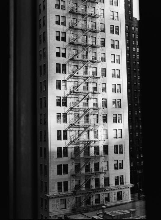 A tall building with many windows and fire escapes is visible from a high vantage point, with another building in the background.